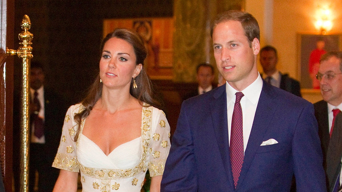 Prince William in a dark blue suit walking next to Kate Middleton in a white and gold gown during their trip to Southeast Asia.