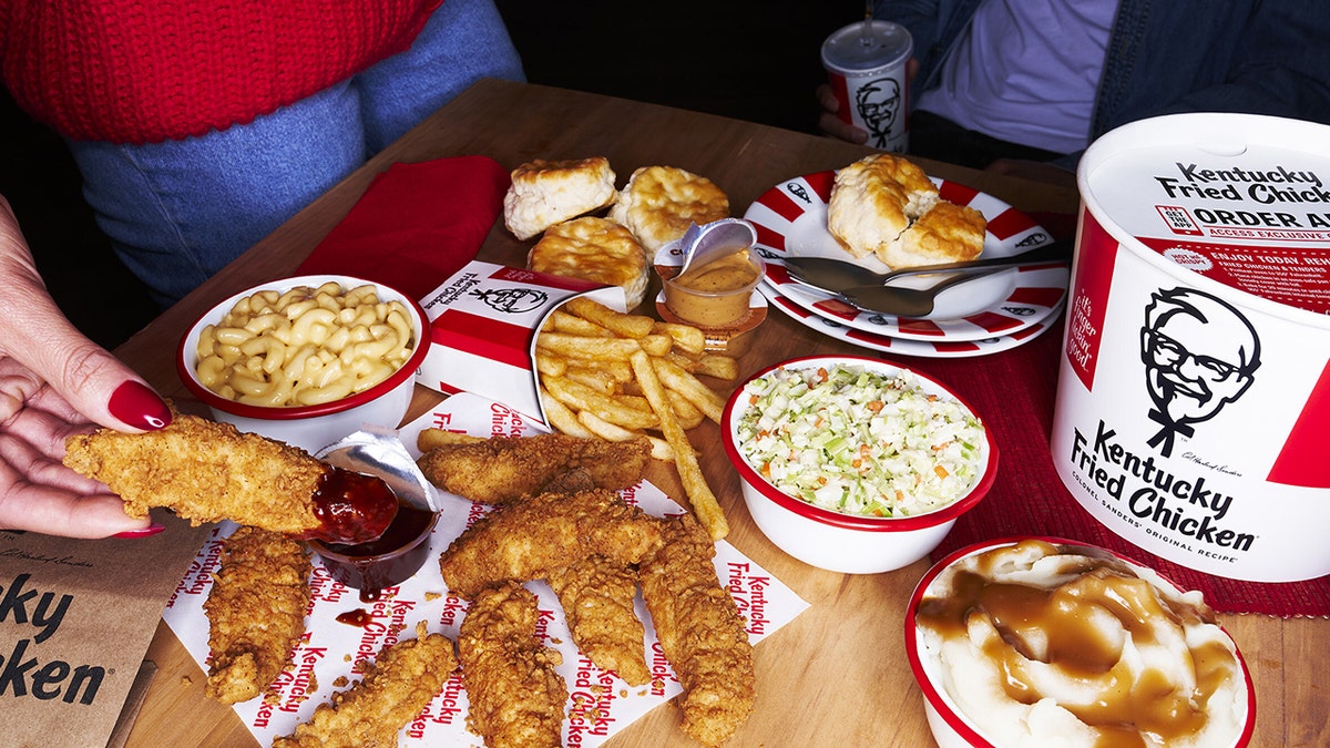 Spread of KFC items including chicken tenders, fries and biscuits and sides from Build-A-Bucket meal seen spread out on table as people reach for it.