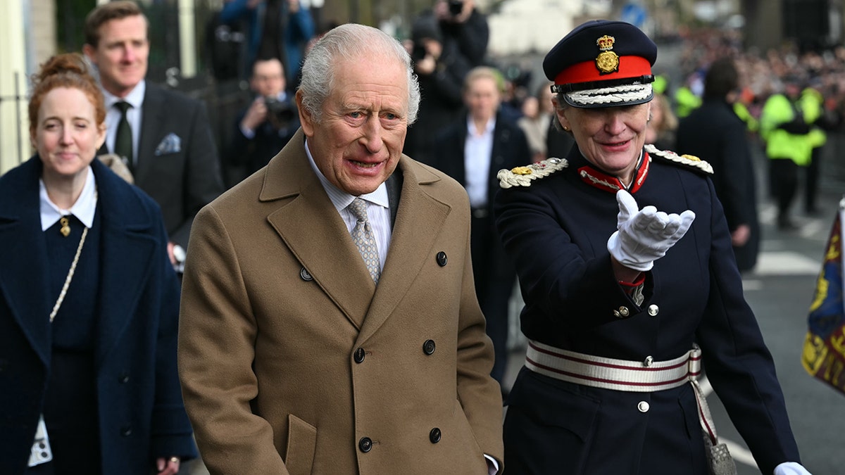 King Charles in a tanned coat walking alongside a royal guard outdoors.