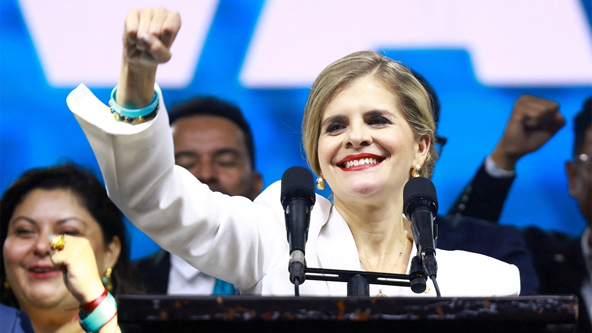 A woman stands on a stage with supporters behind her, responding to early election results during a campaign event.