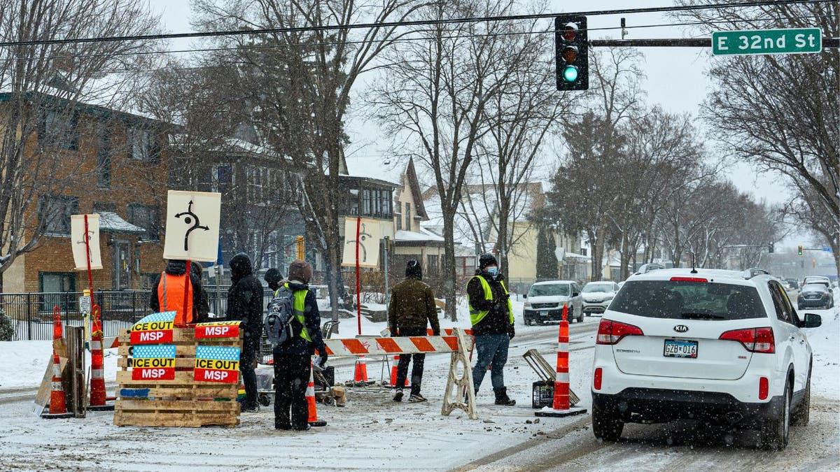 Masked people stand by a wooden barricade with signs while vehicles wait on a snowy street.