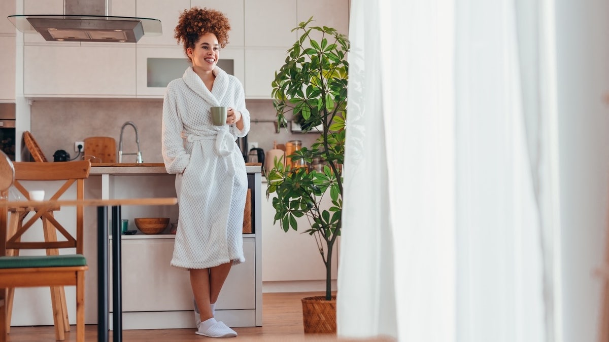 woman wearing bathrobe drinking coffee in kitchen looking out window