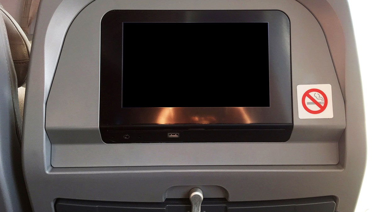 Airplane seatback entertainment screen with USB port and no smoking sign inside a commercial aircraft cabin.