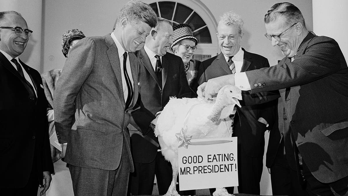 President Kennedy smiling as he admires a 55-pound turkey, which wears a sign reading "Good Eating, Mr. President!" outside the White House.