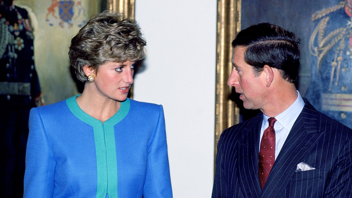 Princess Diana in a blue and green suit in a serious conversation with Prince Charles in a dark blue suit.