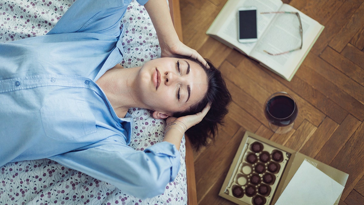 Girl lying on her bed with her eyes closed and thinking, book, phone and glasses on floor next to glass of red wine and box of chocolate
