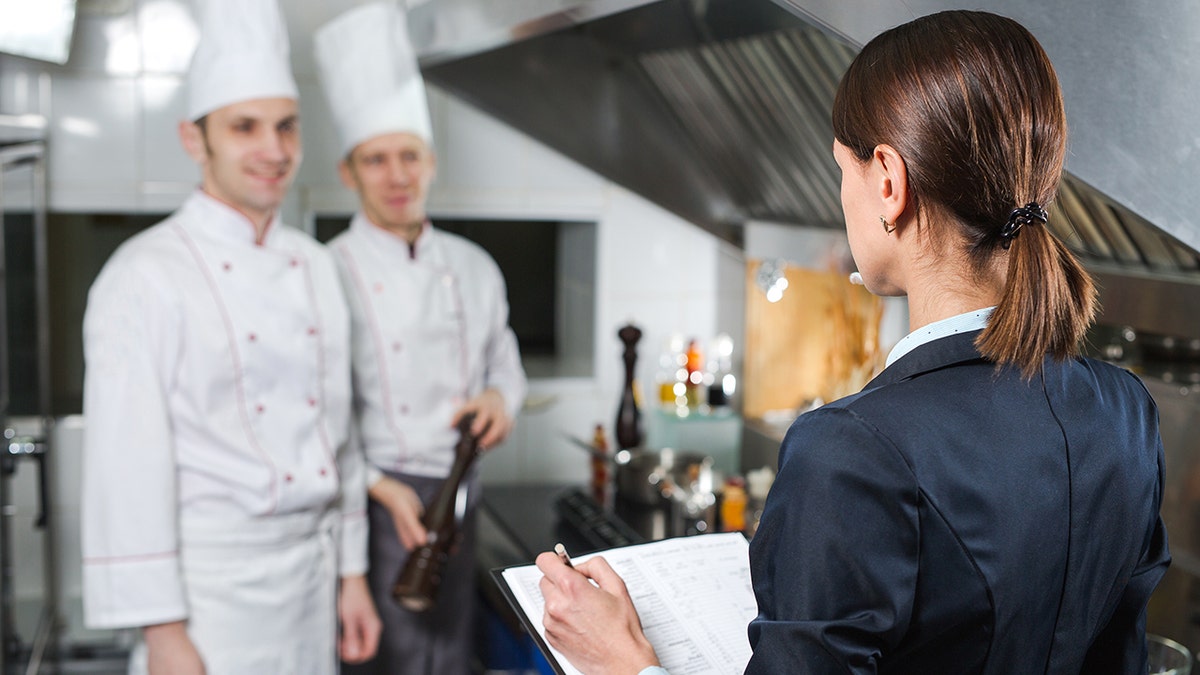 Woman with clipboard conducts check at restaurant in kitchen, seen with two chefs