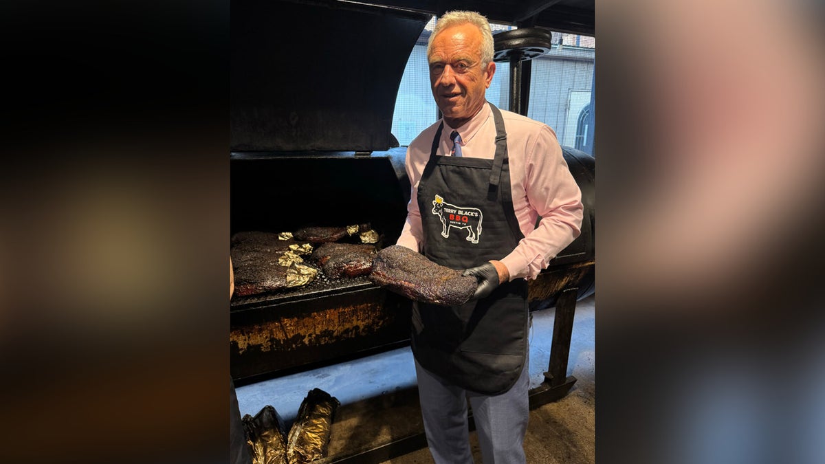 HHS Secretary RFK Jr. hold rack of ribs in front of smoker at Terry Black's BBQ in Augtin Texas. Kennedy seen smiling with pink button down, gloves and black Terry Black's apron.