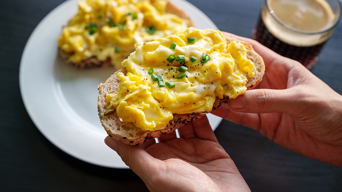 A woman's hand holding a piece of toasted bread topped with scrambled eggs.