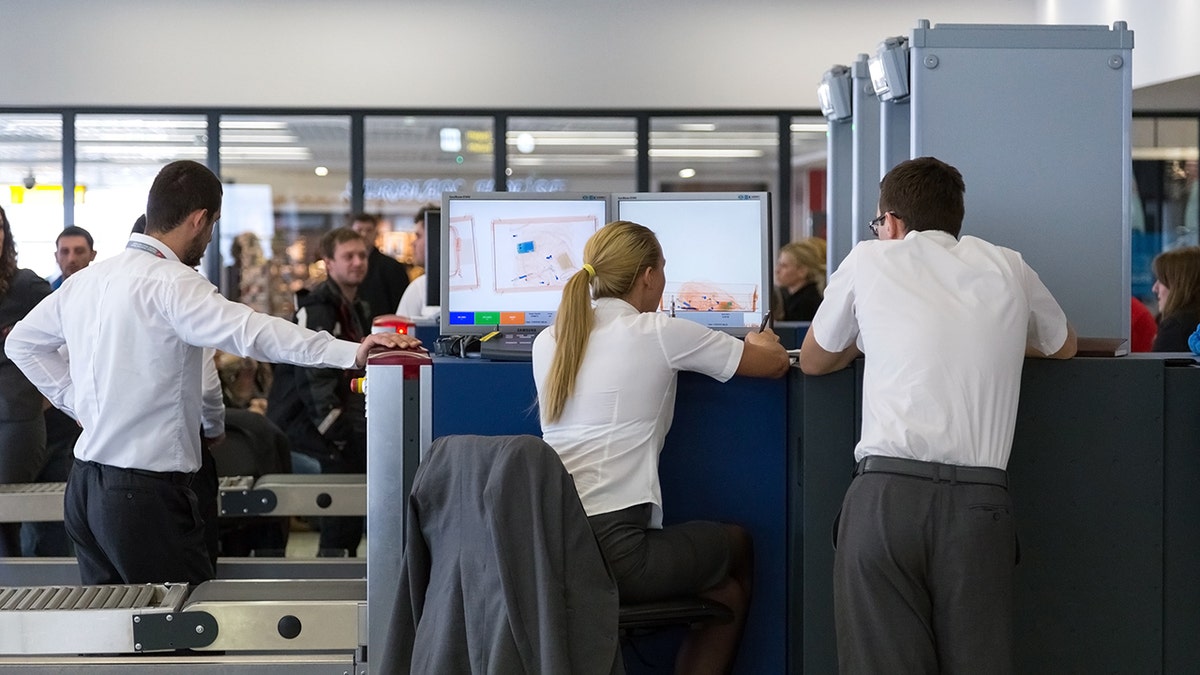 Airport security officers review X-ray scanner images at a TSA checkpoint while monitoring passenger luggage.