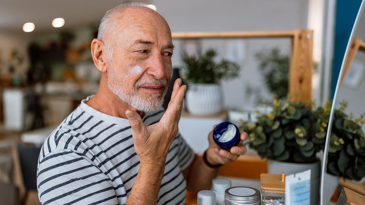 Handsome bearded senior man applying cream at his face and looking at himself with smile while standing in front of the mirror. Self care concept.