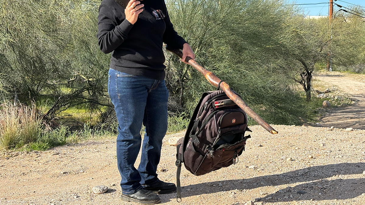 A woman holds up a black SwissGear brand backpack with a walking stick in the desert of Tucson.