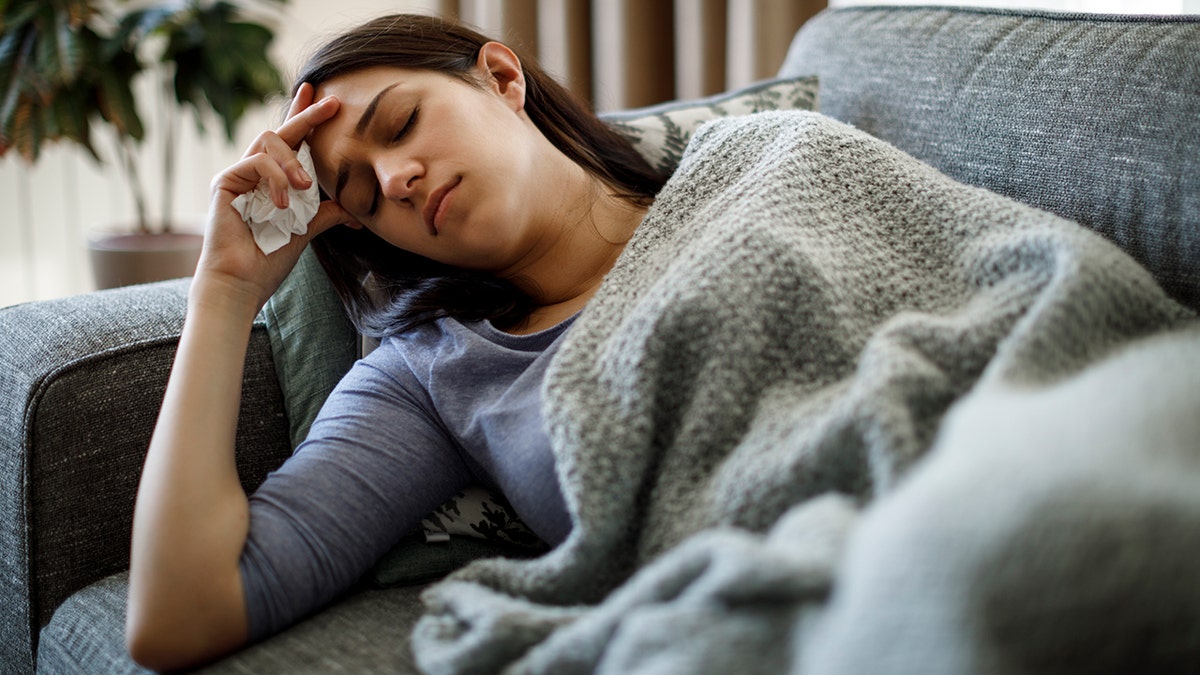Woman resting on a couch with a blanket, holding a tissue to her head and appearing fatigued at home.