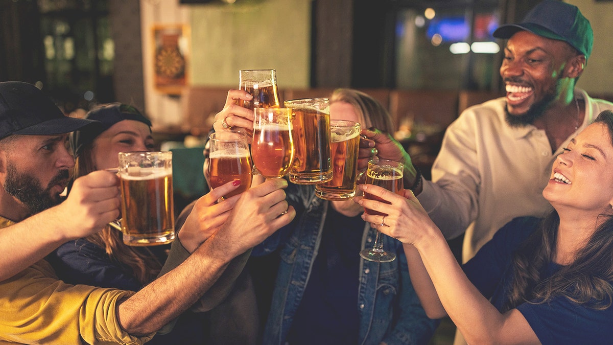 Young people hold beer glasses as they cheers while standing in a bar.