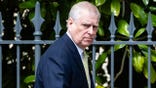 Ex-Prince Andrew looking annoyed in a dark blue suit and yellow tie walking in front of a fence.