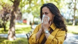 Portrait of unhealthy cute  female in yellow top with napkin blowing nose, looks to the source of the allergy, place for advertising.