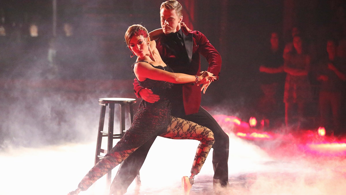 Amy Purdy and Derek Hough perform an intense ballroom dance routine on a smoky stage, posed in a dramatic lunge under red lighting.