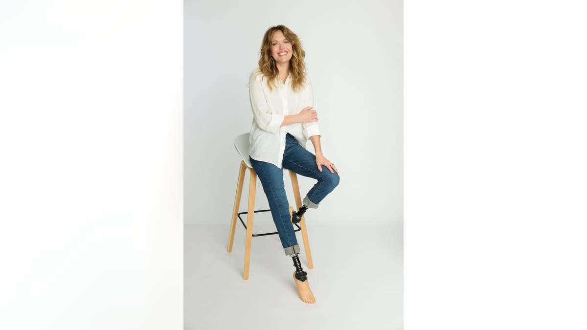 Amy Purdy sits on a tall wooden stool wearing a white blouse and rolled-up jeans, smiling, with her prosthetic legs visible against a white studio background.
