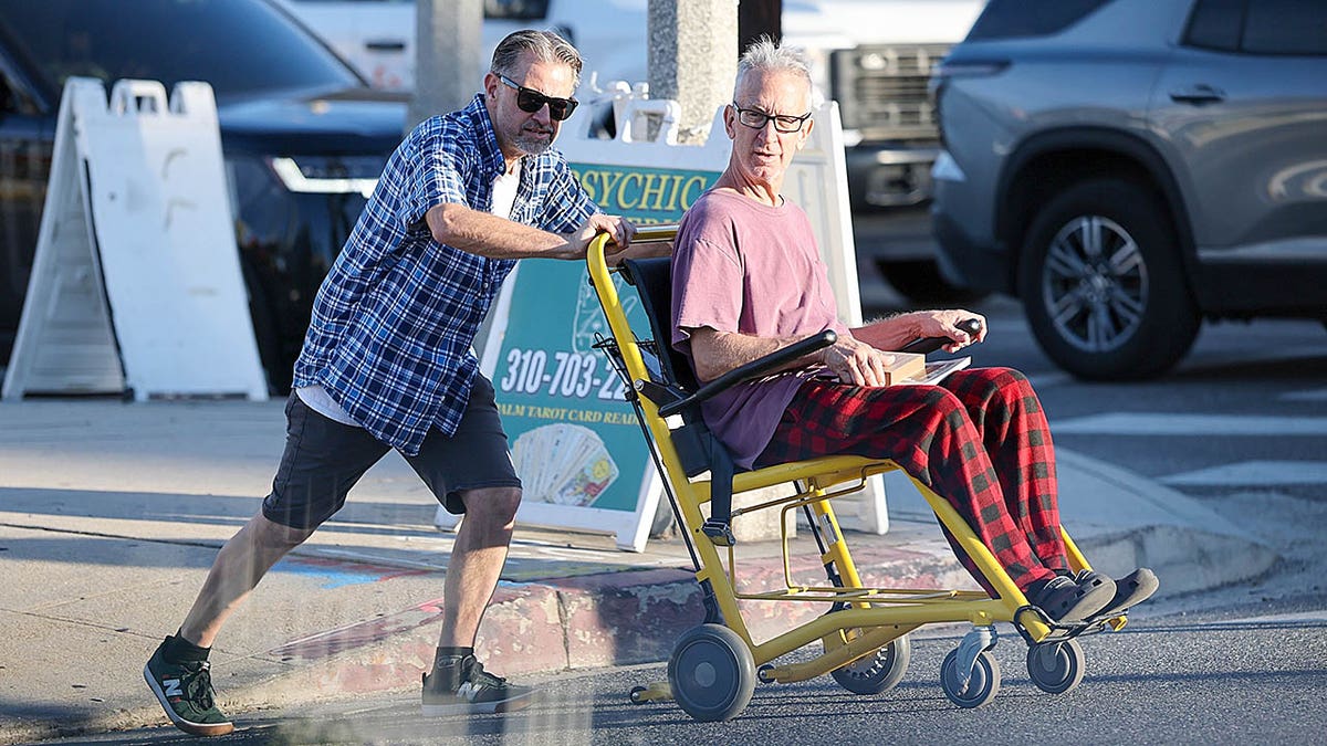 Andy Dick sits in a wheelchair as a man pushes him across a street