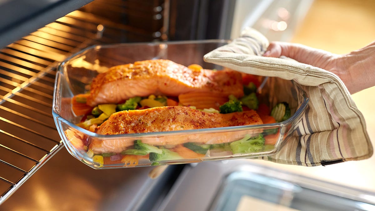 Woman with potholder taking baking dish with salmon fish and vegetables out of oven at home kitchen.