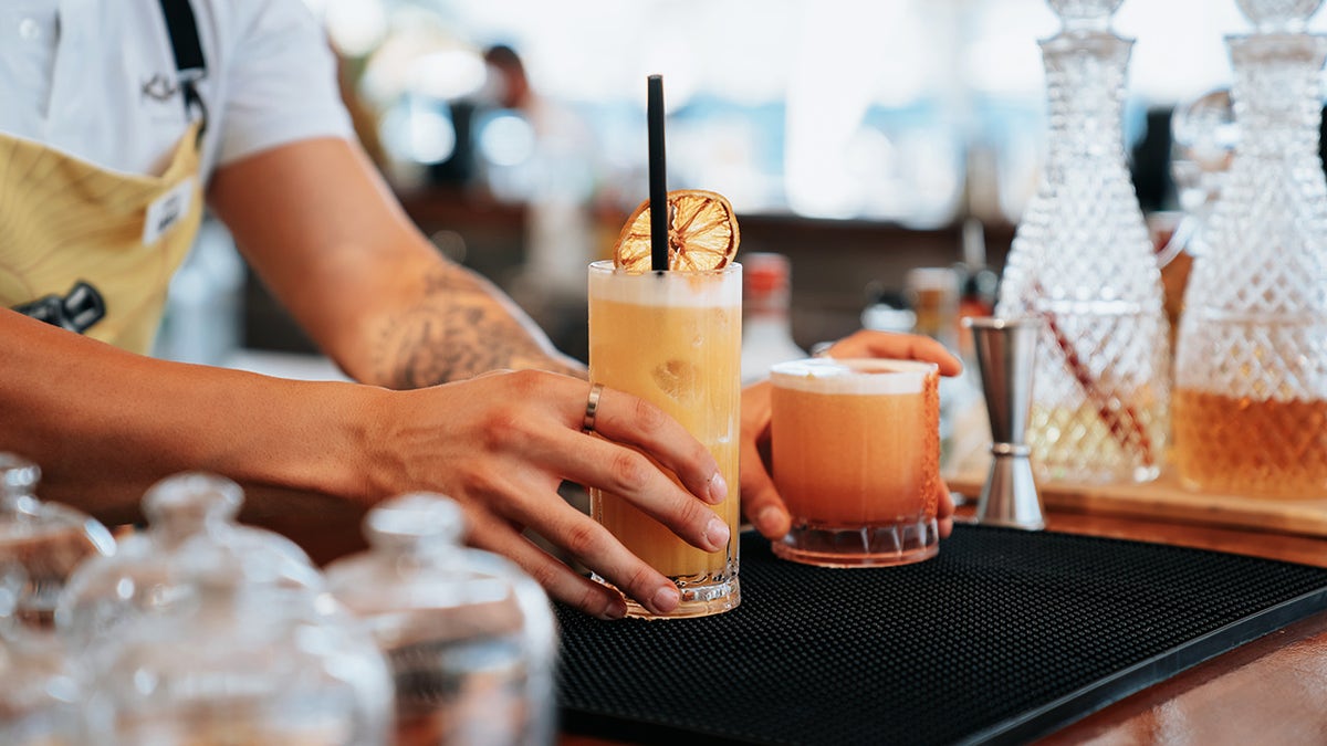bartenders hands seen placing two craft cicktails onto bar, garnished with dehydrated fruits, crystal decanters seen around bar.