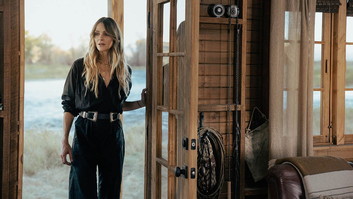 Beau Garrett in a black outfit standing in the doorway of a rustic wooden cabin near a lake.