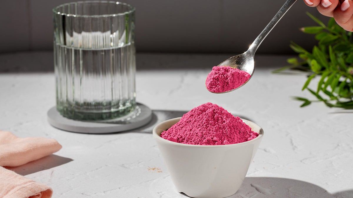 Beetroot powder being scooped out of small white bowl with a spoon and glass of water in background.