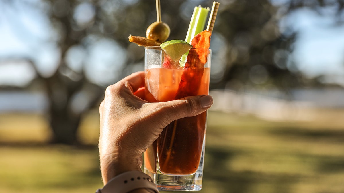 A hand toasts a Bloody Mary at a tranquil waterway