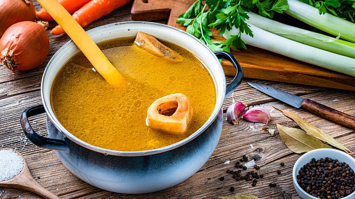High angle view of a cooking pan filled with homemade bone broth shot on rustic wooden table. Ingredients for cooking bone broth are all around the pan