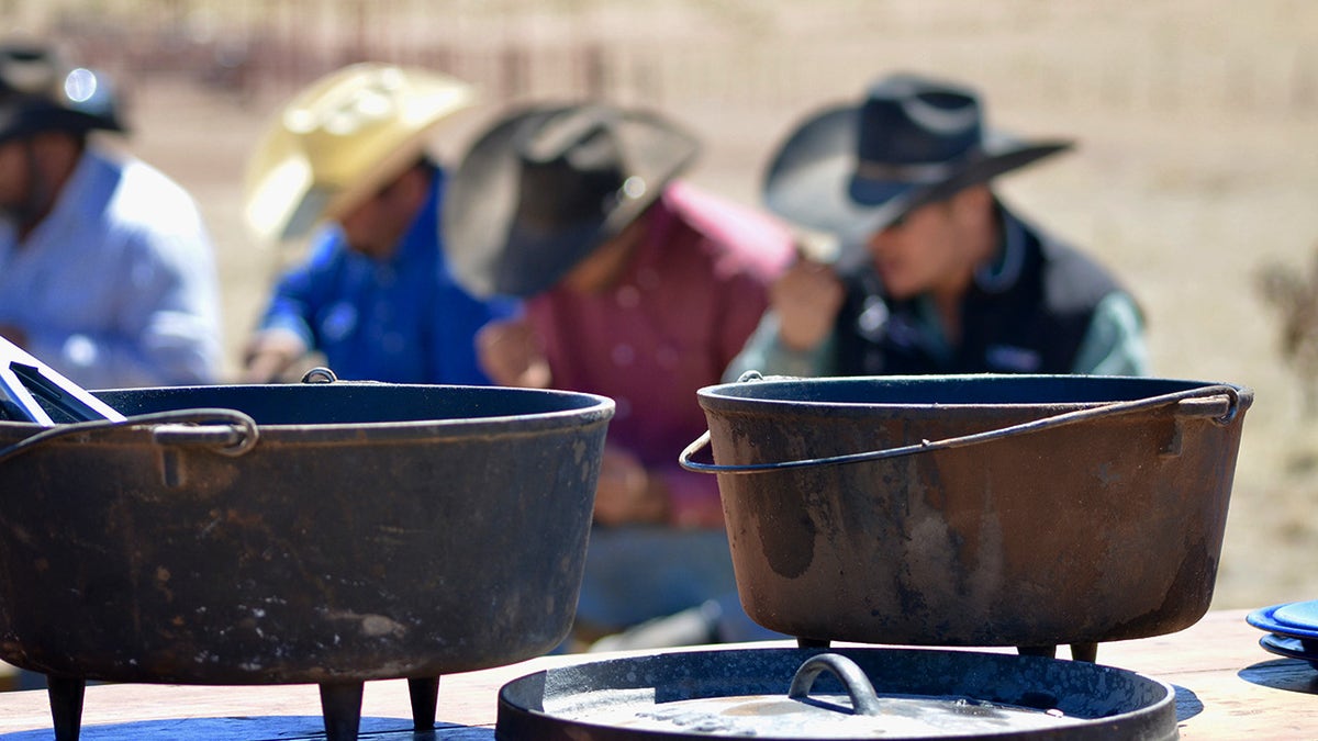 Cowboys eating meal on frontier, seen behind pots on burners.