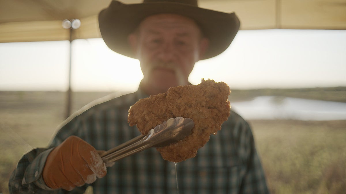 Cowboy chef Kent Rollins holding chicken-fried steak with tongs at campsite on the frontier.
