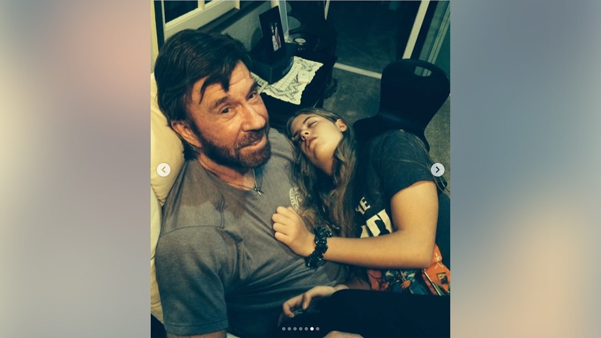 Chuck Norris looking at the camera with his daughter sleeping on his lap.