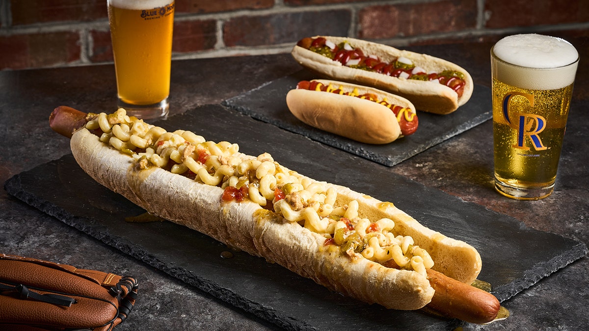 Two-foot, one-pound hot dog topped with mac and cheese and other toppings surrounded by beers and other foods at baseball stadium.