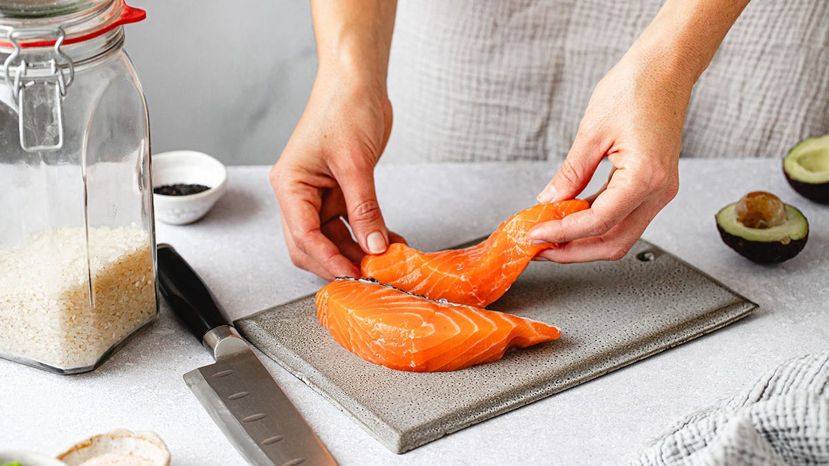 Woman's hands seen prepping raw salmon on cutting board with rice, knife and avocado around her.