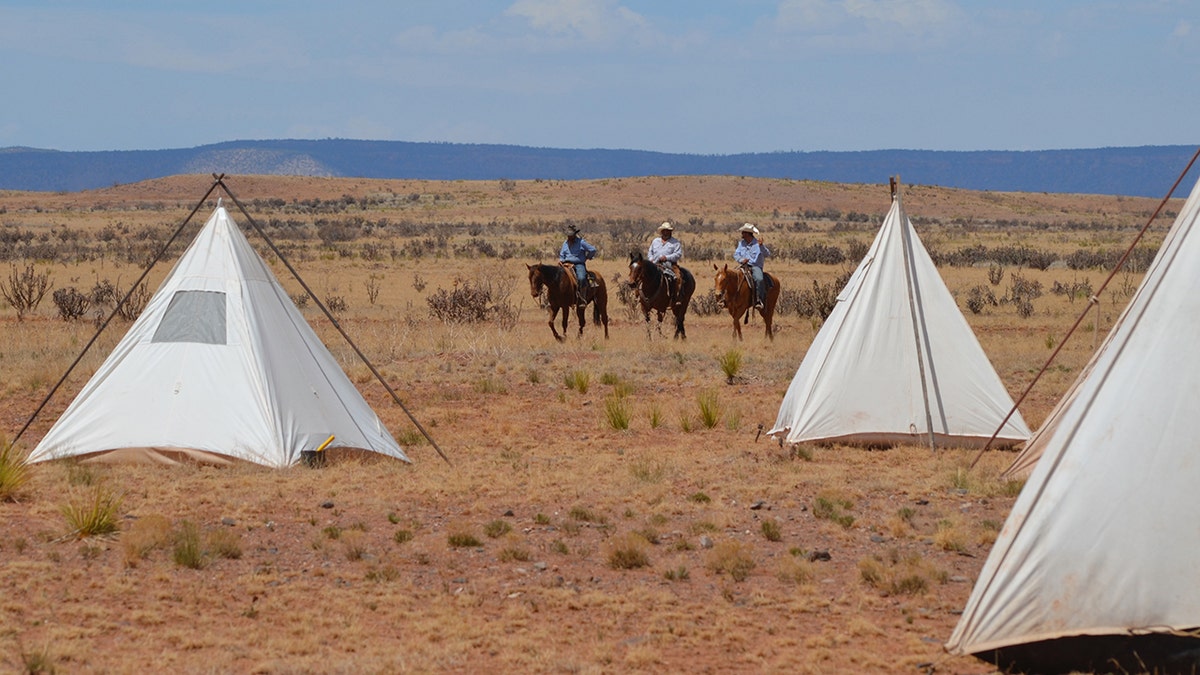 Cowboys on horseback riding toward tents with mountains in background.