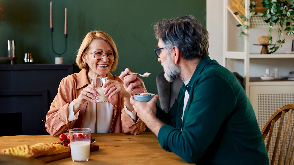 An elderly woman drinks milk from a glass while her better half eats cereal as they sit together at the breakfast table.