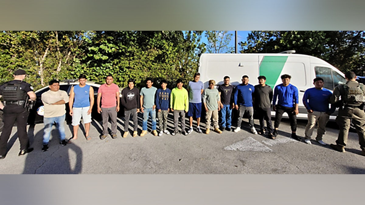 Fourteen individuals are lined up facing the camera between two men in uniform with their backs to the camera.