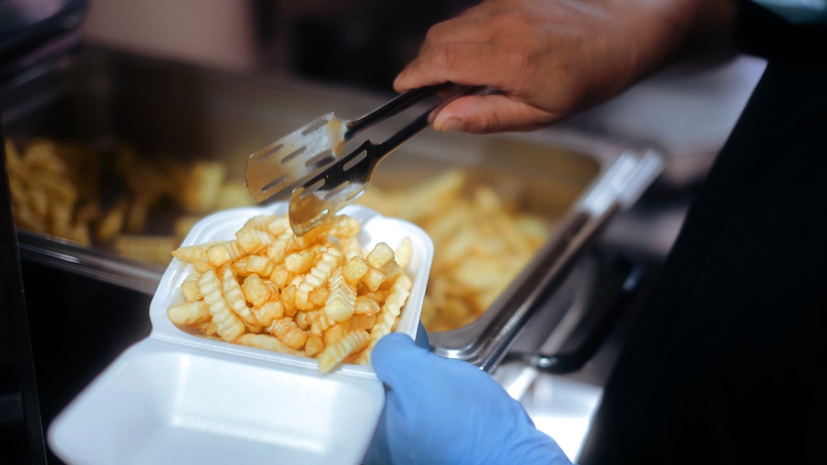 Professional Cook Putting French Fries in a Plastic Food Container; Restaurant worker filling a polystyrene with a takeaway order.