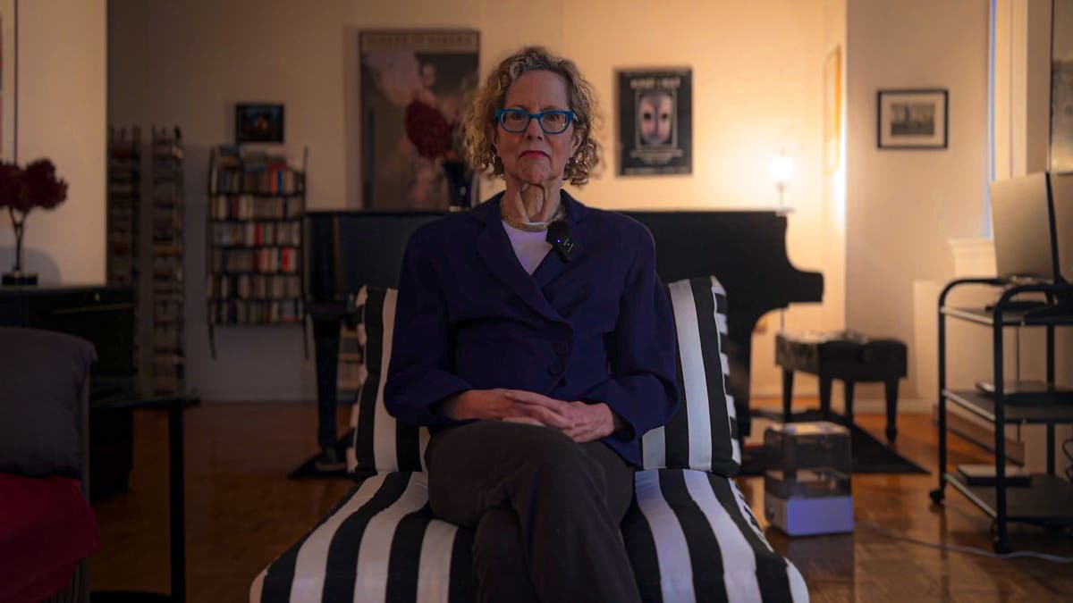 Blond, curly haired white woman with black rimmed glasses sitting on sofa in center of living room