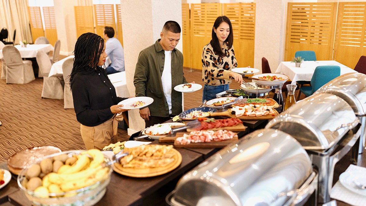 Three people at the hotel breakfast buffet table preparing food for themselves.