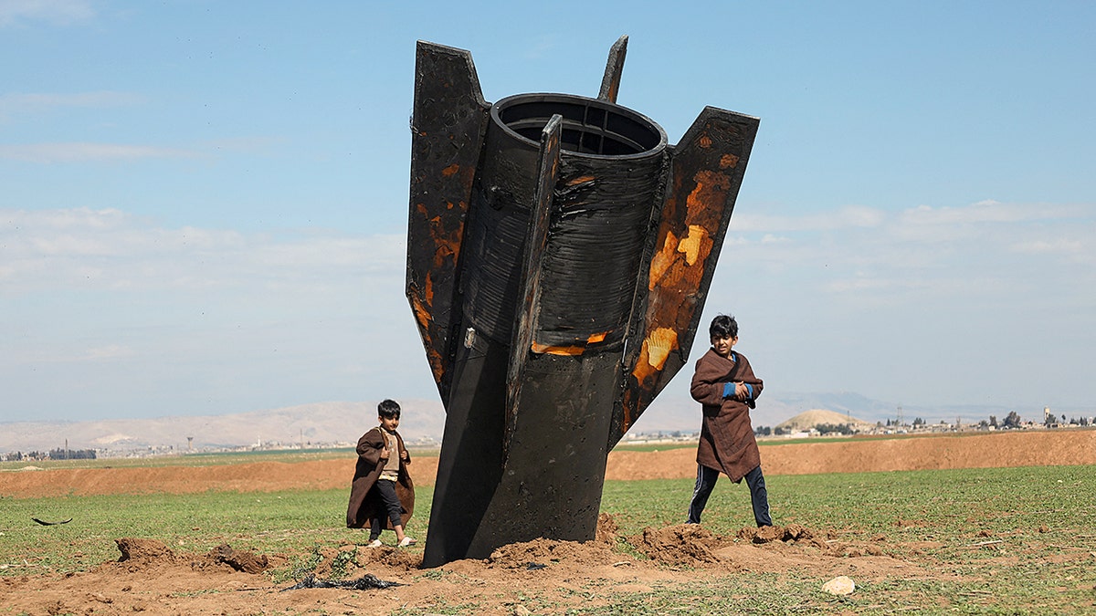 Kids walking around a large downed Iranian missile in a dirt field.