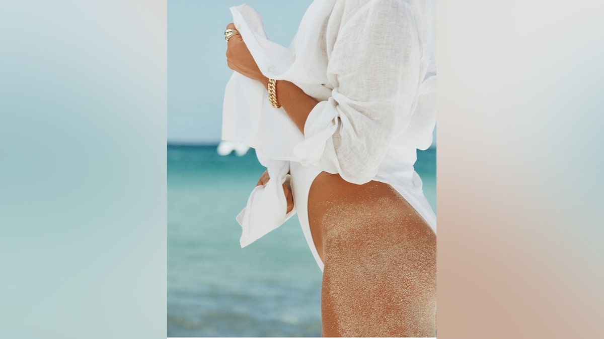 Kristin Cavallari standing on the beach in a white cover-up with sand on her leg.