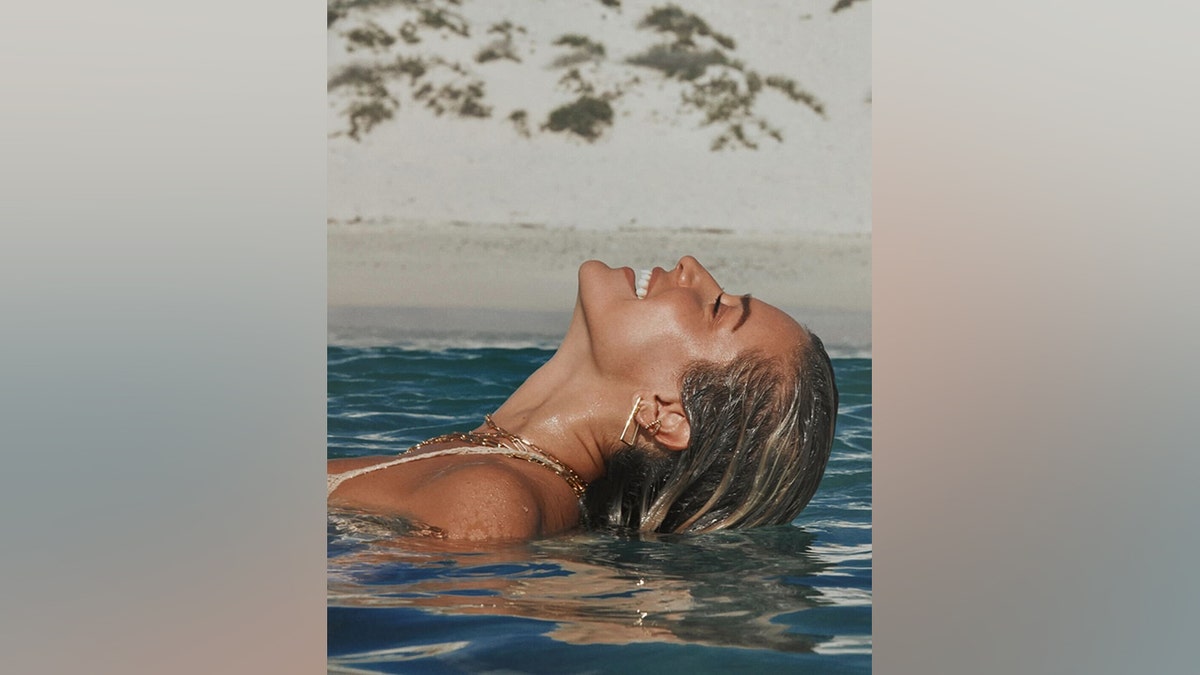 Kristin Cavallari floats in the ocean with her head tilted back and eyes closed, wearing gold necklaces and earrings.