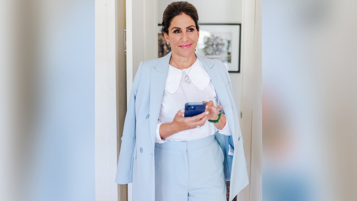Lisa Mirza Grotts, a certified etiquette expert, standing smiling in light blue suit holding phone.