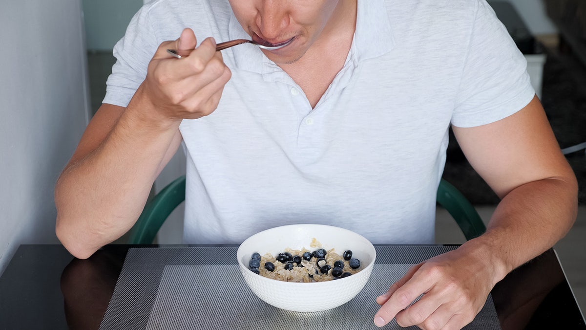 Man in white t-shirt eats delicious porridge cereals with blueberries and milk for breakfast close up