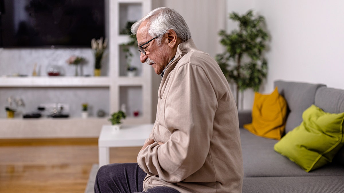 An older man sitting on a sofa holding his abdomen in pain.