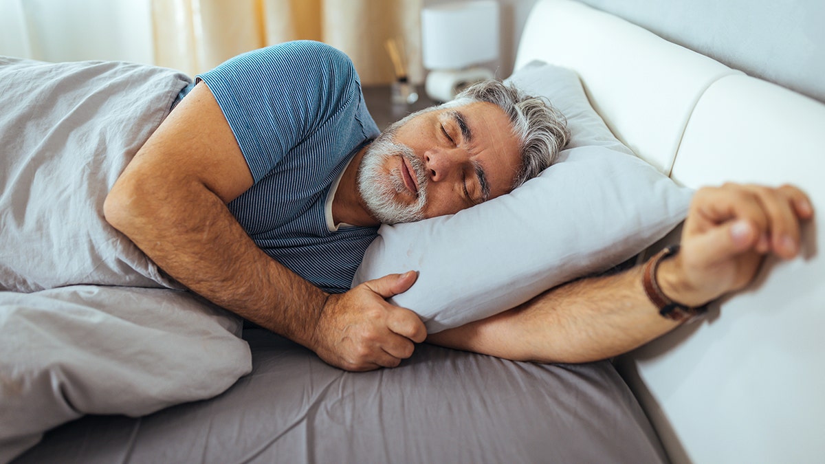 Middle-aged man sleeping on his side, hugging a white pillow in a bright, comfortable bedroom.