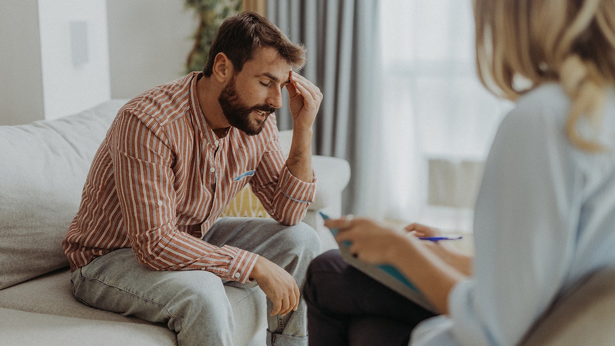 man sits on couch with hand on forehead while talking with a female therapist