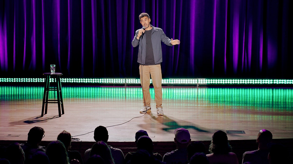 Mark Normand stands onstage holding a microphone during his stand-up comedy special.