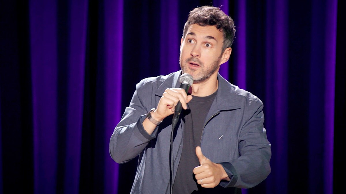 Mark Normand stands onstage holding a microphone during his stand-up comedy special.